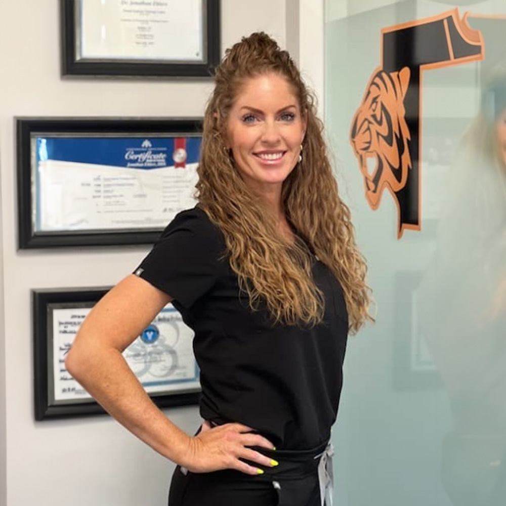 A smiling woman wearing a black shirt with a badge on her waist, standing in front of a framed diploma and a wall with a logo, posing for the camera.