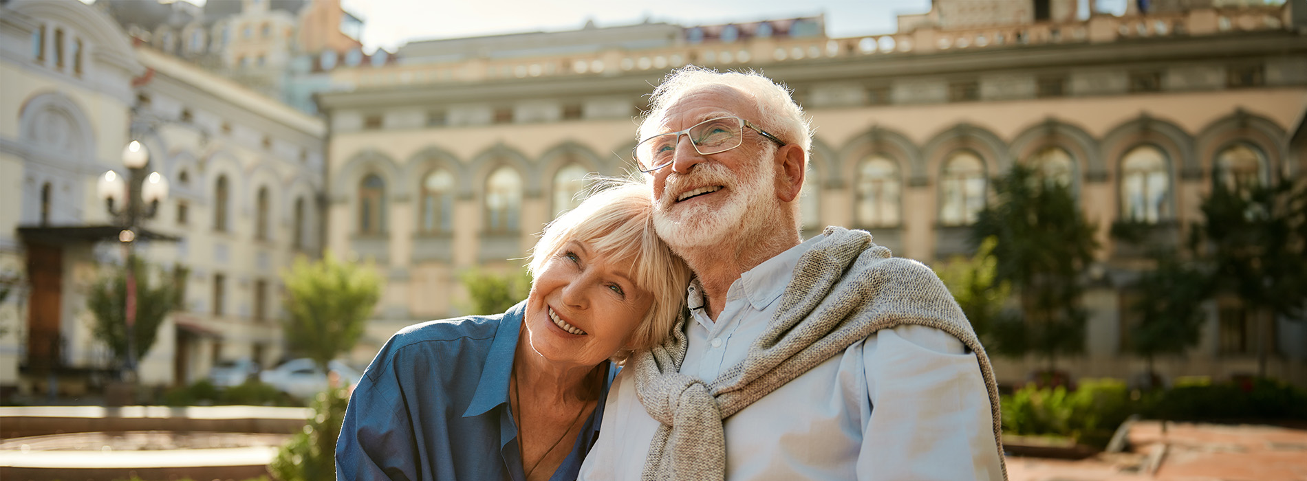 An elderly couple poses together outdoors with a cityscape in the background.