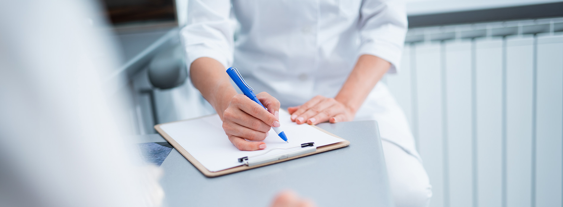A person writing on a clipboard at a desk with a blurred background.