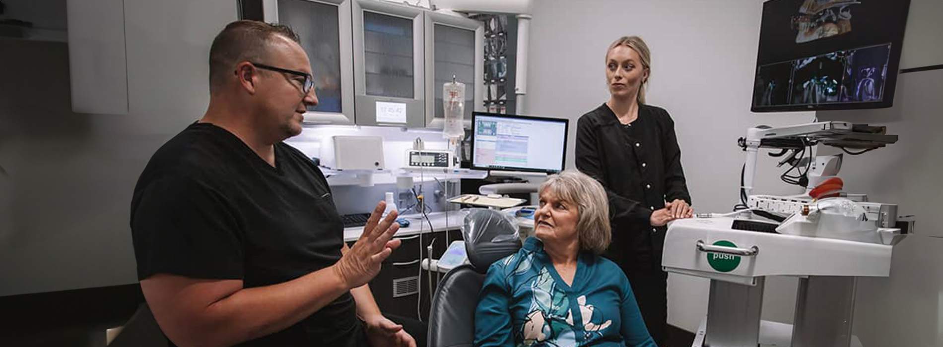 A person is seated in a dental chair, receiving care from a dental professional who stands behind them.