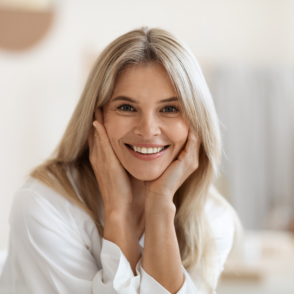 The image shows a woman with blonde hair smiling at the camera while holding her hand on her face. She has fair skin and appears to be wearing makeup. The background is blurred but suggests an indoor setting, possibly a room with a mirror.