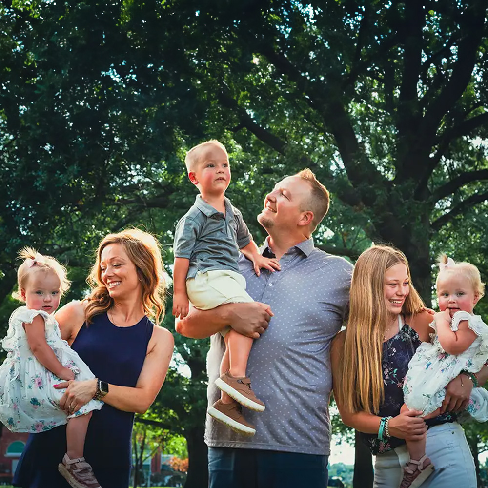 A family posing together with their children outdoors during daylight man holding two young children while standing between a woman and another adult, all smiling and looking at the camera.