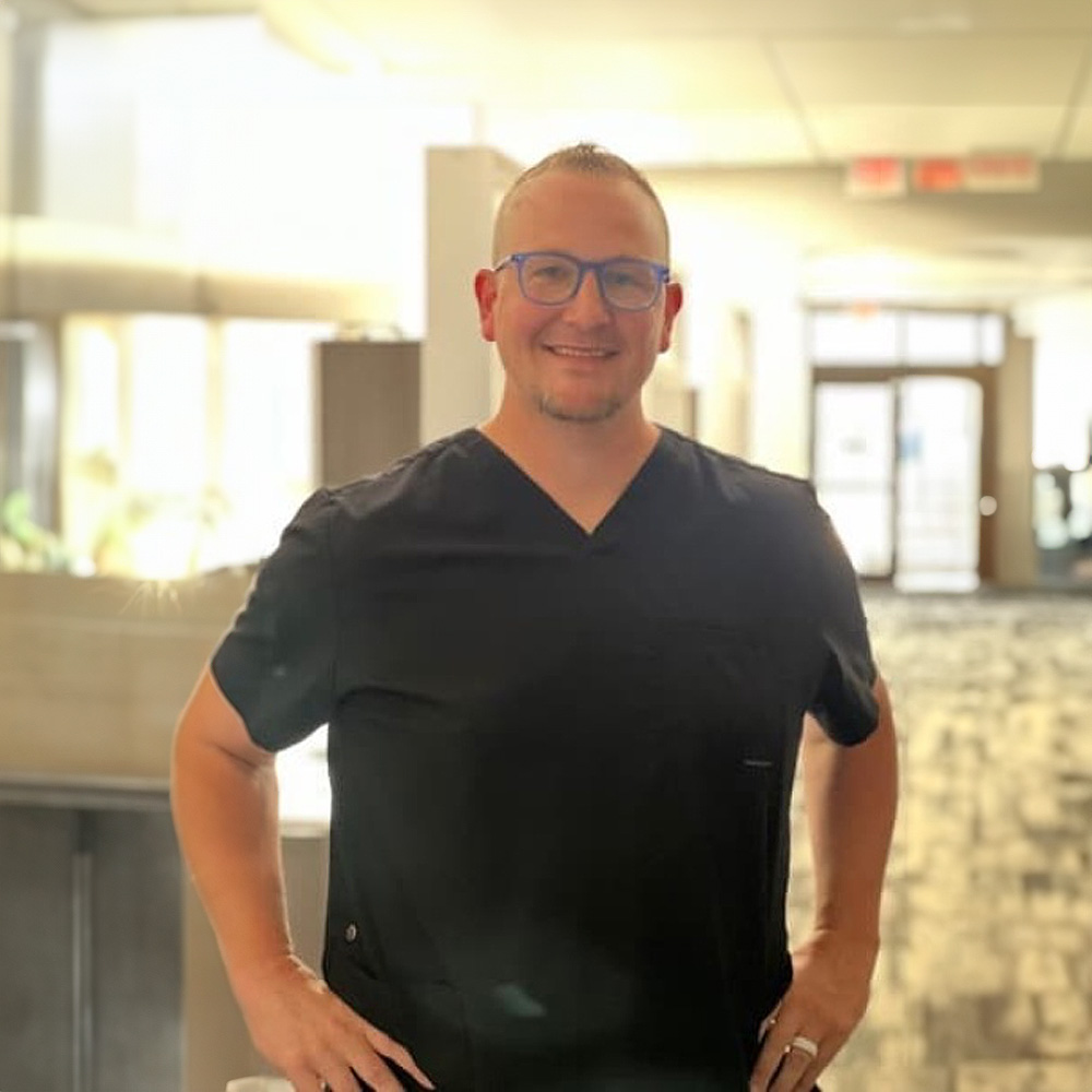 The image shows a man wearing glasses, standing indoors with his arms crossed, posing for a portrait.