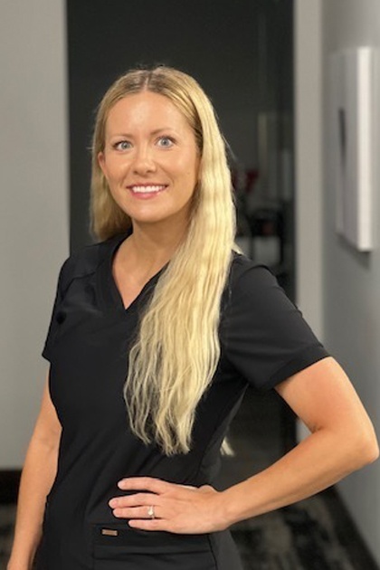 The image features a woman with long blonde hair wearing a dark top and posing for a portrait with a neutral background, likely taken at a professional setting such as an office or workplace.