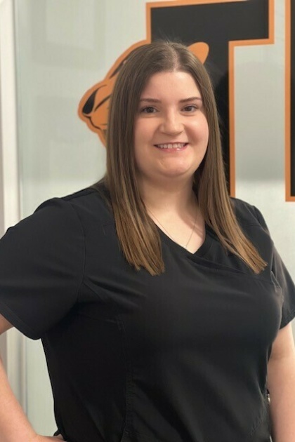 A woman wearing a black shirt stands in front of a wall with a logo, smiling at the camera.
