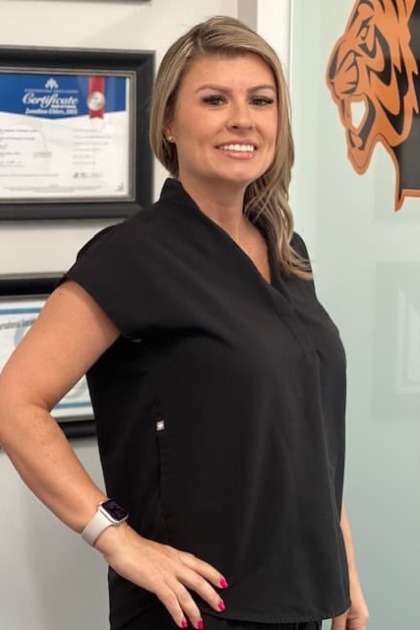 This is a photograph of a woman standing in an office environment, wearing a black blouse and dark pants, posing for the camera with a smile.