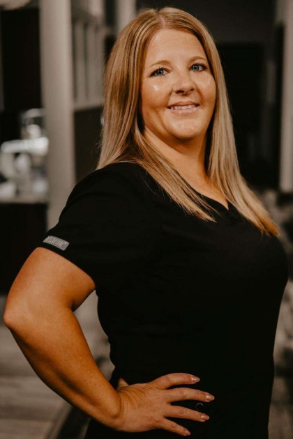 Woman in black shirt and white pants posing with hands on hips against dark background.