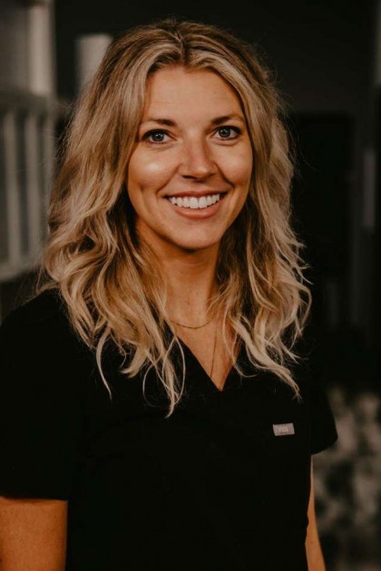 A woman with long hair, wearing a black shirt, stands smiling against a dark background.
