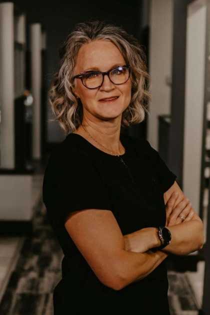 A woman with short hair, wearing glasses, stands confidently with her arms crossed in front of her, against a blurred background featuring mirrors.