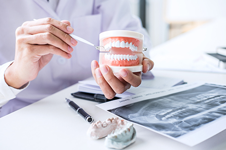 Image 1 A dental professional holding a tray with a model set of teeth and viewing it through a magnifying glass, with a background showing medical equipment and a skeletal model. Image 2 The same dental professional holding the same model set of teeth in front of a display of dental implants and tools, with an X-ray image visible in the background.