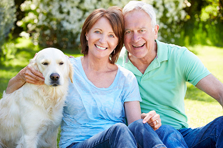 The image shows an elderly couple sitting outdoors with a golden retriever dog on their laps, smiling at the camera.